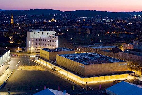 Luftaufnahme des BERNEXPO-Geländes, explizit der neuen Festhalle bei Dämmerung, mit Blick Richtung Stadtzentrum und sichtbarem Münster und Verwaltungsbauten.